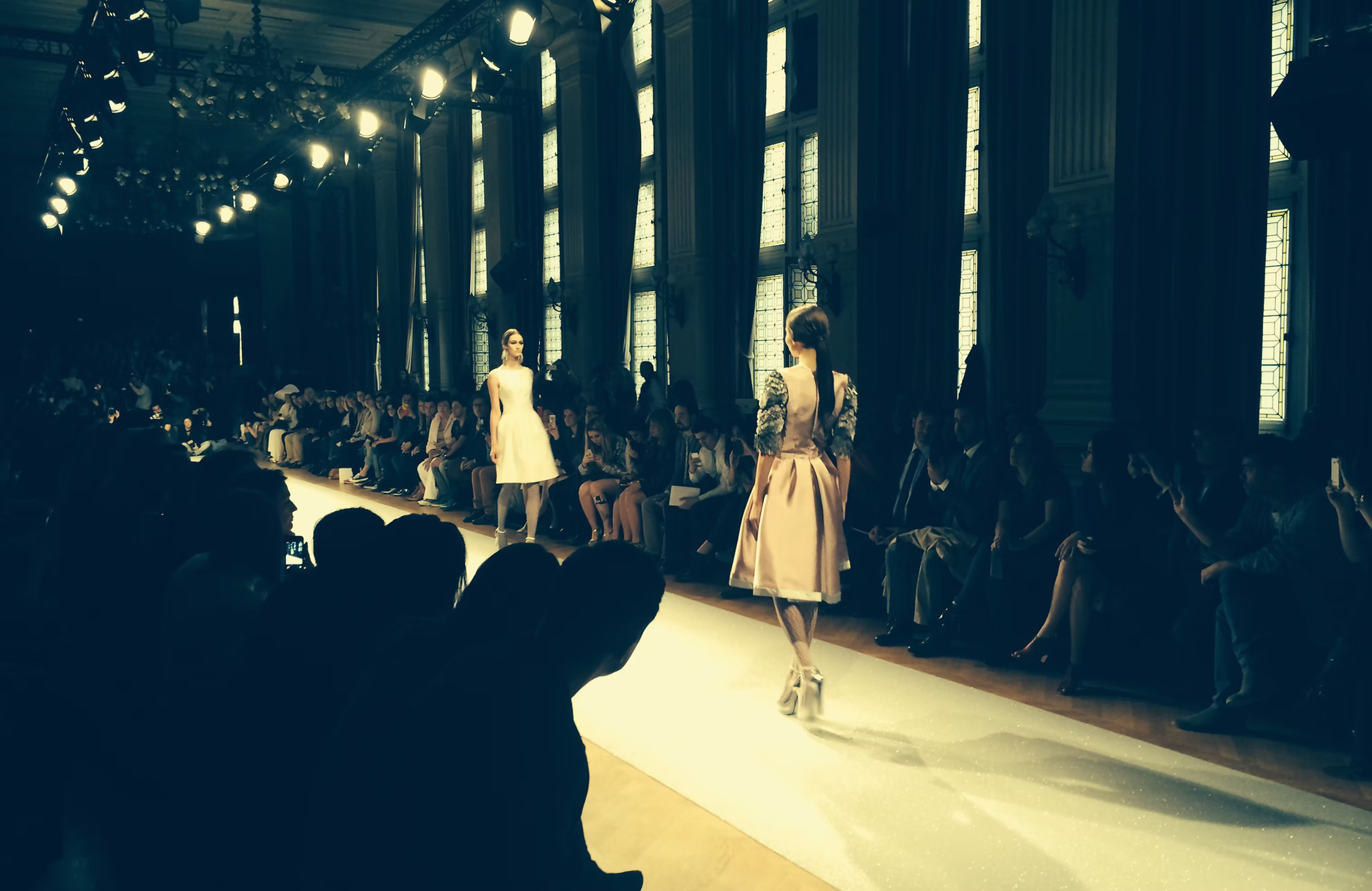 Models walking an illuminated runway in a hall with tall windows
